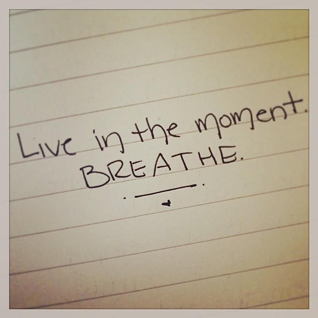 A handwritten spiritual inspirational quote on lined paper saying "live in the moment. breathe." with a small heart doodle underneath.