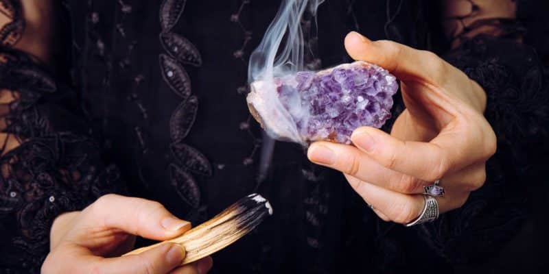 How To Cleanse Crystals - 11 Powerful Ways To Charge Crystals 12 A woman's hand using palo santo to smoke cleanse an amethyst crystal cluster.