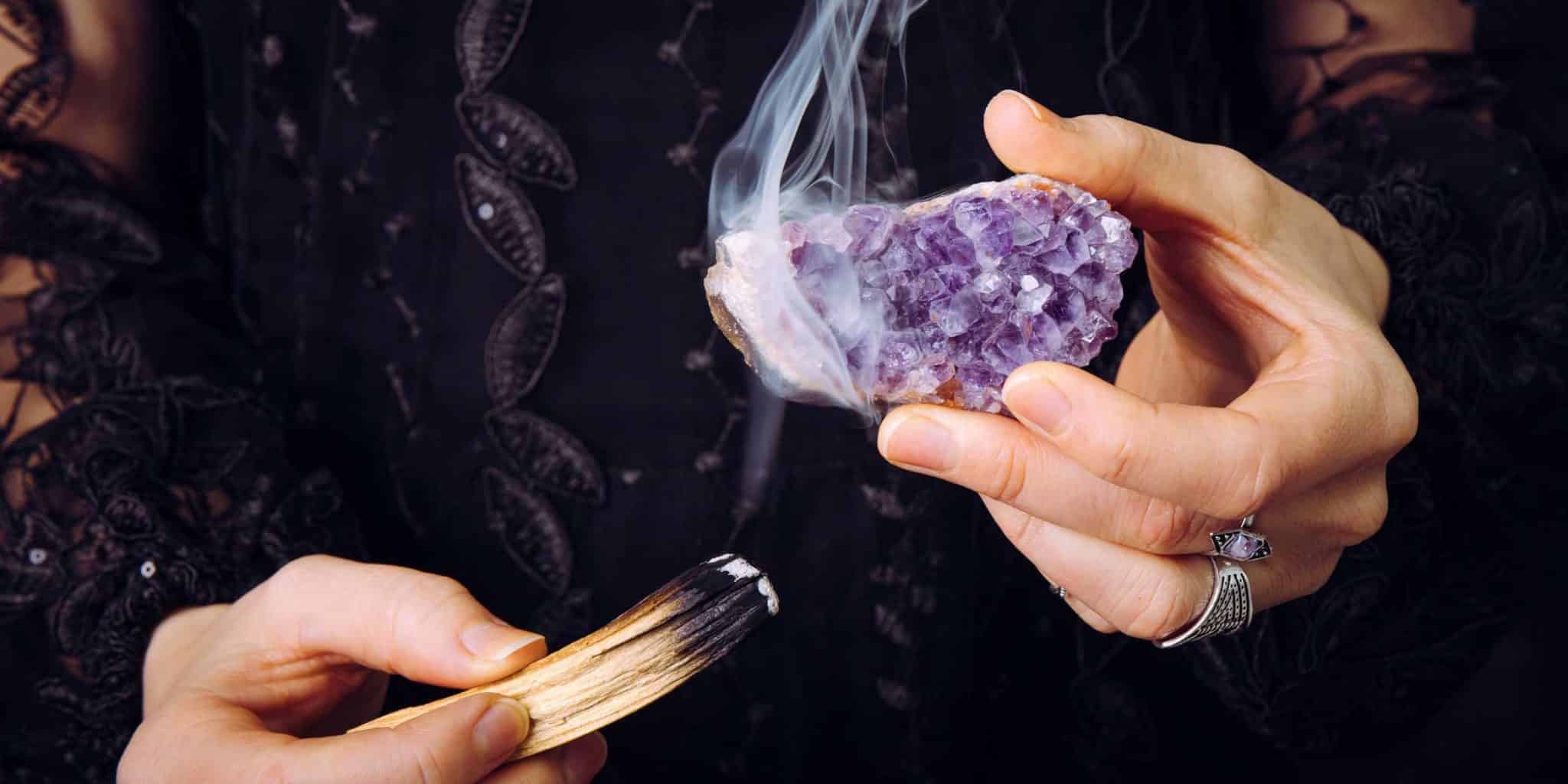 A woman's hand using palo santo to smoke cleanse an amethyst crystal cluster.