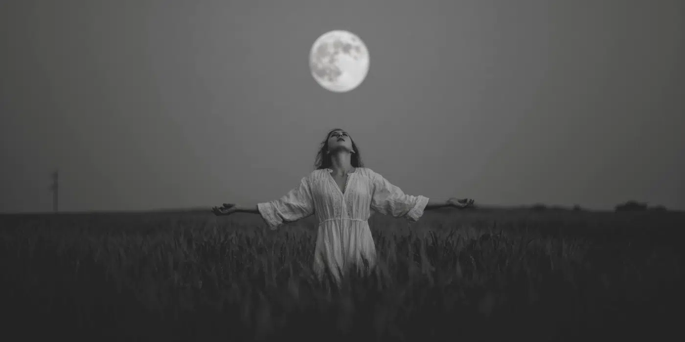 A woman standing under a full moon with her arms outstretched as she does a full moon chant