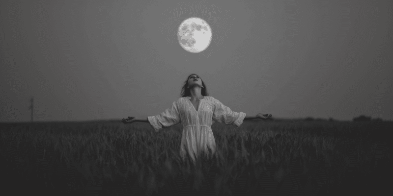 A woman standing under a full moon with her arms outstretched as she does a full moon chant