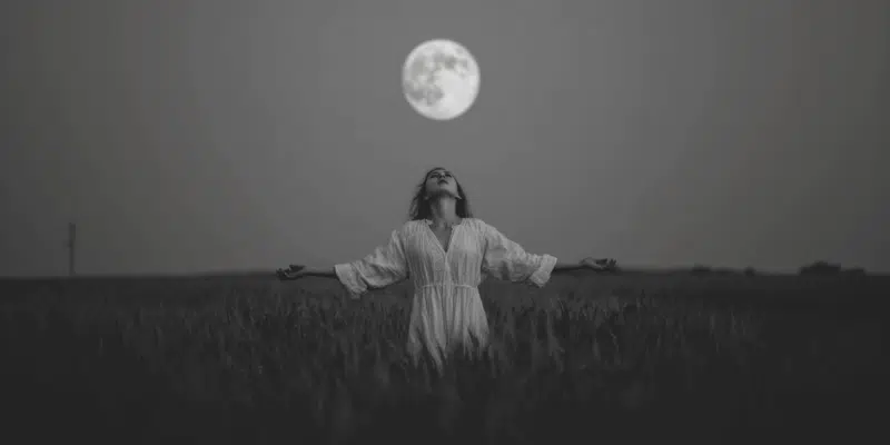 A woman standing under a full moon with her arms outstretched as she does a full moon chant