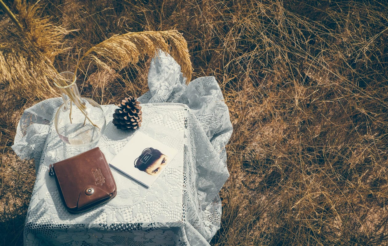 Rustic outdoor tranquility captured with a vintage wallet, a pocket mirror, and a pine cone artfully placed on a lace-draped wooden crate amidst golden wild grass, creating an almost witch