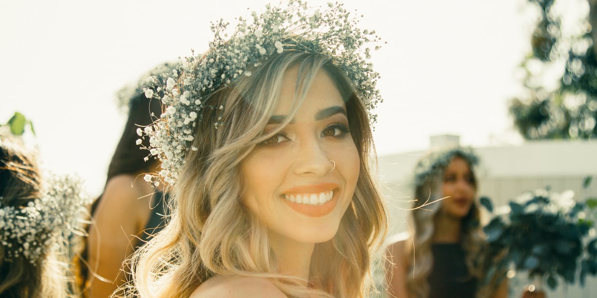 A radiant woman with a witchy floral crown smiles on a sunny day, surrounded by others in a joyful and festive atmosphere.