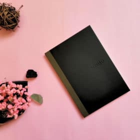 Free Materia Magicka Worksheets 3 A minimalistic black notebook with the word "miniso" embossed on the cover, presented on a plain background beside a bowl of pink flowers and a small nest-like ball, designed as a new