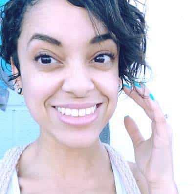 A cheerful woman with a bright smile, wavy hair, and sparkling eyes, playfully posing for a happy, casual selfie with an occult charm hanging around her neck.