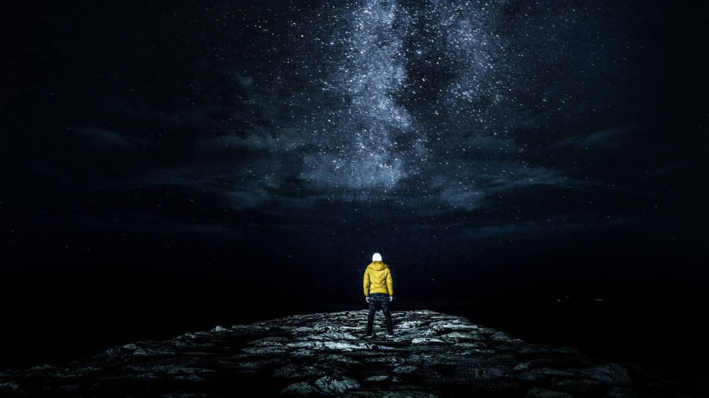 A lone adventurer in a yellow jacket stands on a rocky outcrop, gazing up at a magnificent star-studded night sky, embracing the vastness of the universe and its metaphysical mysteries.