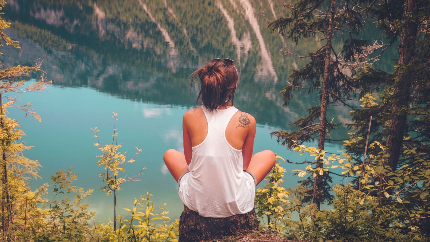 A person sitting on a rock, gazing out at a tranquil turquoise lake surrounded by forested mountains, feeling a deep spiritual connection to nature.