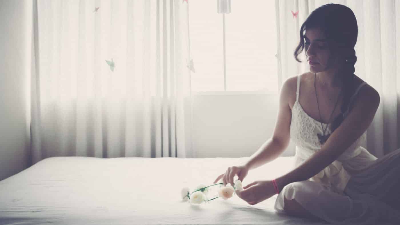 A contemplative young woman sitting by a window, gracefully touching a stem of white flowers on a bed, bathed in soft, witchy light.