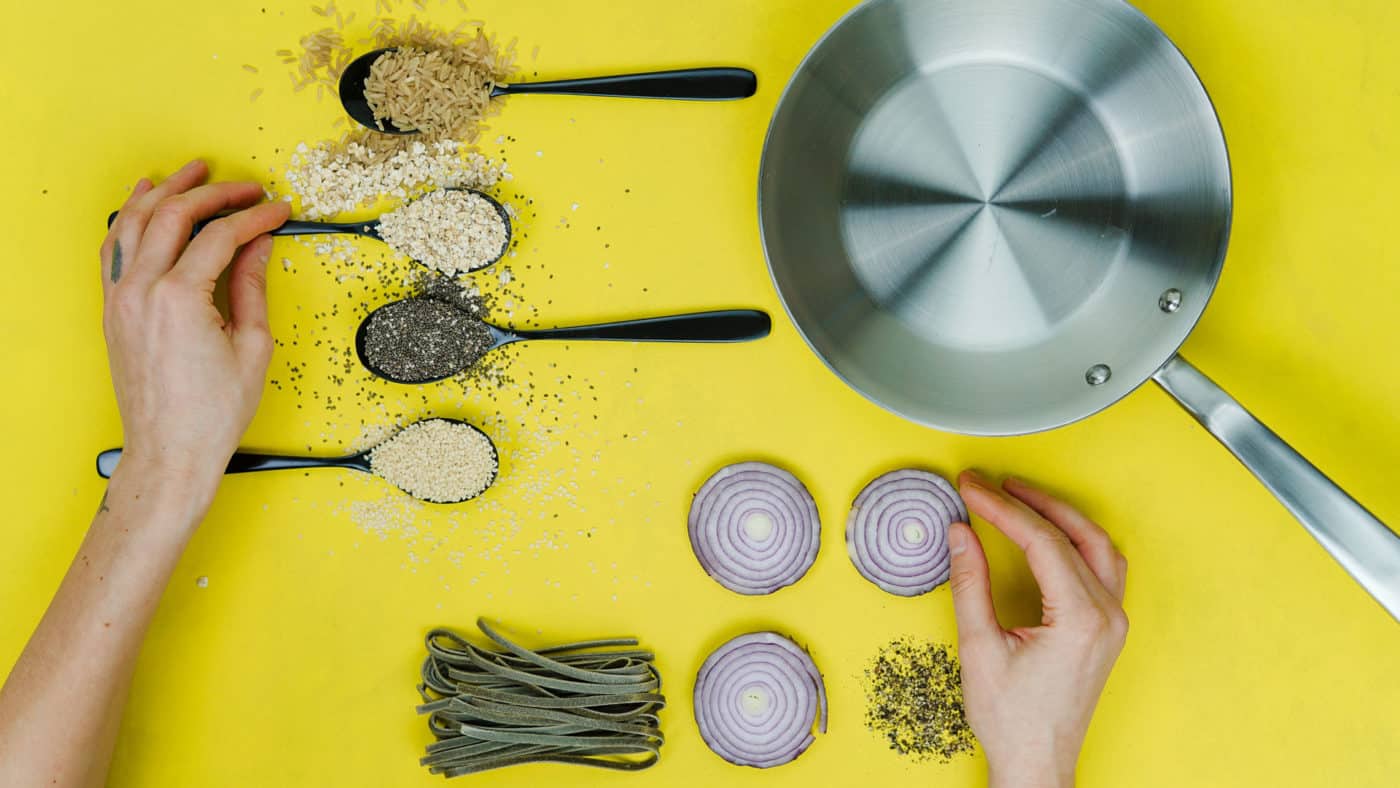 A colorful cooking preparation scene with a variety of grains and pasta measured in spoons, slices of red onion arranged symmetrically, and a person's hands adding a touch of seasoning against a vibrant yellow background