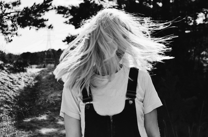 A black-and-white photograph capturing a spontaneous moment where a woman's hair is playfully tossed by the wind, obscuring her face as she stands on a path amid a natural, spiritual setting.