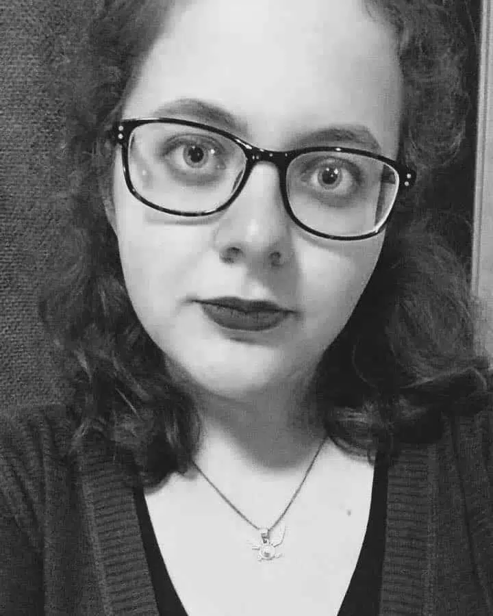 A black and white portrait of a young woman with glasses wearing dark lipstick and a necklace from a metaphysical shop, posing before a plain background.