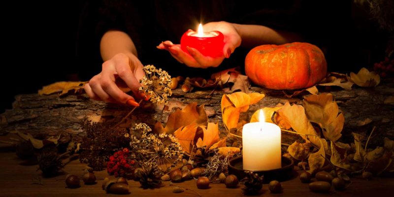 Samhain Ritual: Connect to your Spirit Family for Growth 3 A person arranges witchy elements like dried flowers, leaves, and acorns on a wooden surface, illuminated by the warm glow of candles, with a small pumpkin adding to the seasonal atmosphere.