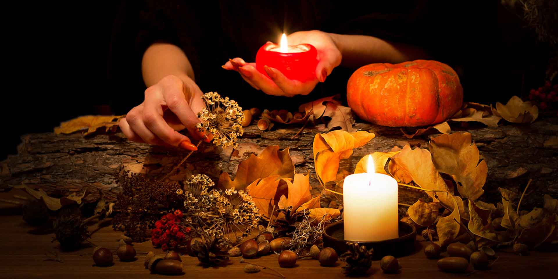 A person arranges witchy elements like dried flowers, leaves, and acorns on a wooden surface, illuminated by the warm glow of candles, with a small pumpkin adding to the seasonal atmosphere.