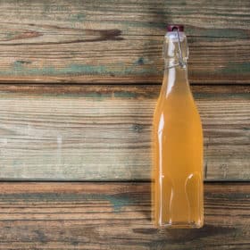 A vintage glass bottle filled with amber-colored liquid secured with a swing-top closure, standing against an aged wooden backdrop with a witchy charm.