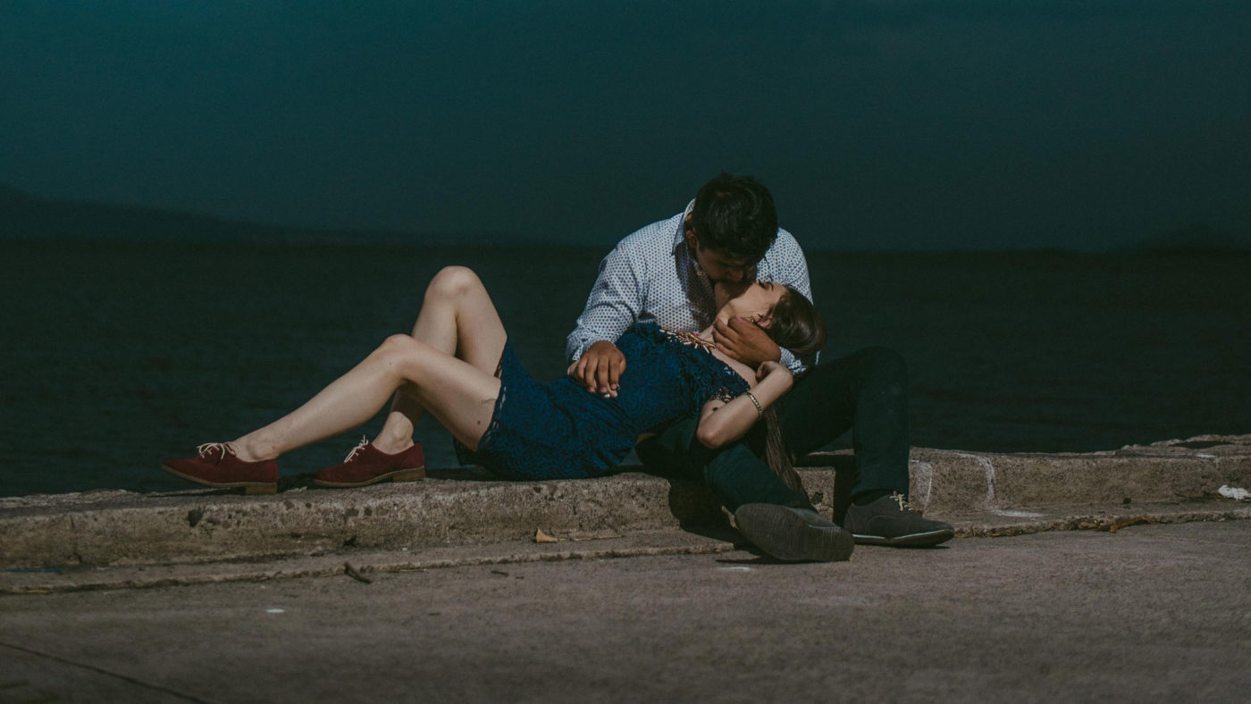 A tender moment by the water: a couple embraces while sitting on a quay under a dusky sky, with a metaphysical shop in the background.