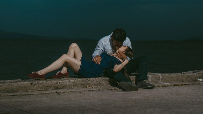 Free Witchcraft Items 3 A tender moment by the water: a couple embraces while sitting on a quay under a dusky sky, with a metaphysical shop in the background.