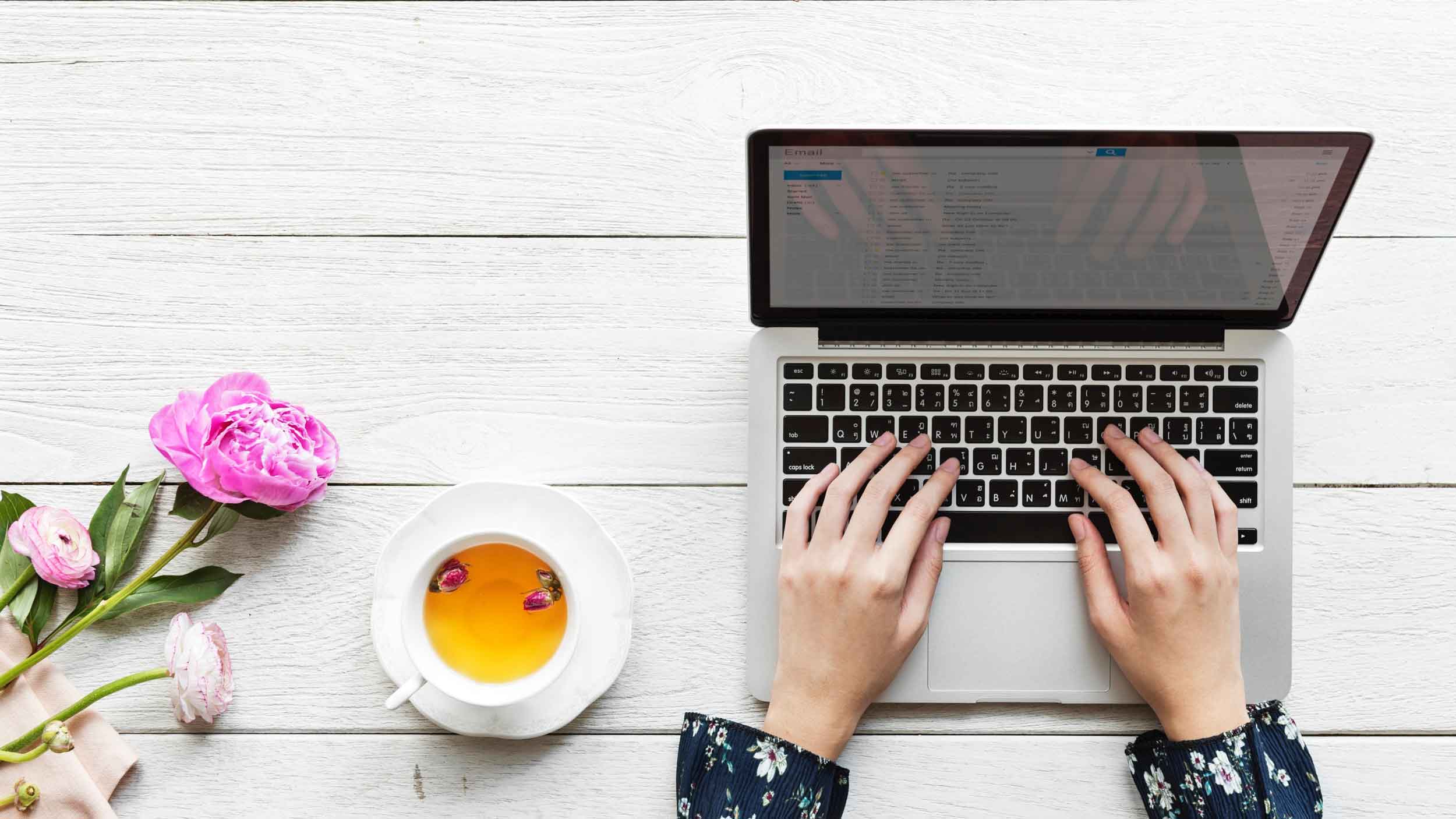 A person working on a laptop with a cup of tea and pink flowers next to a new age product on a white wooden table.