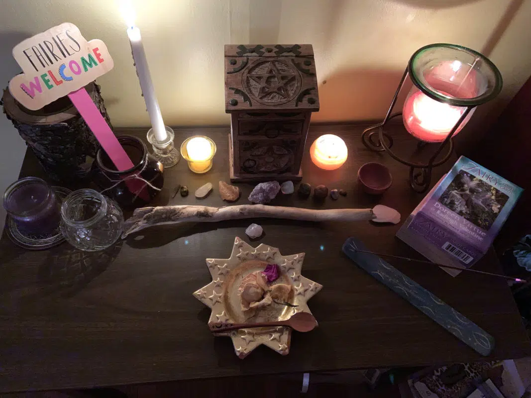 Midsummer: The Reawakening of a Witch 2 Witches Summer Solstice altar with a lit white candle, rose petals, crystals, a wand, and a wooden herb cabinet with a pentacle carved into it.