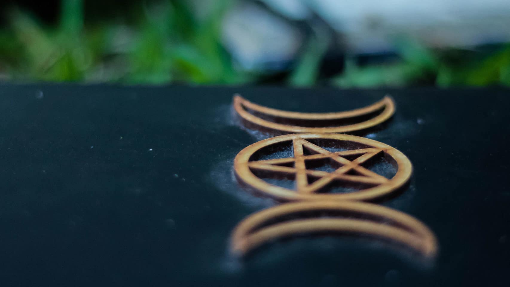 A symbol often associated with the occult and witchy practices, a wooden pentacle interlaced with a crescent moon, rests on a dark surface against a blurry green background.