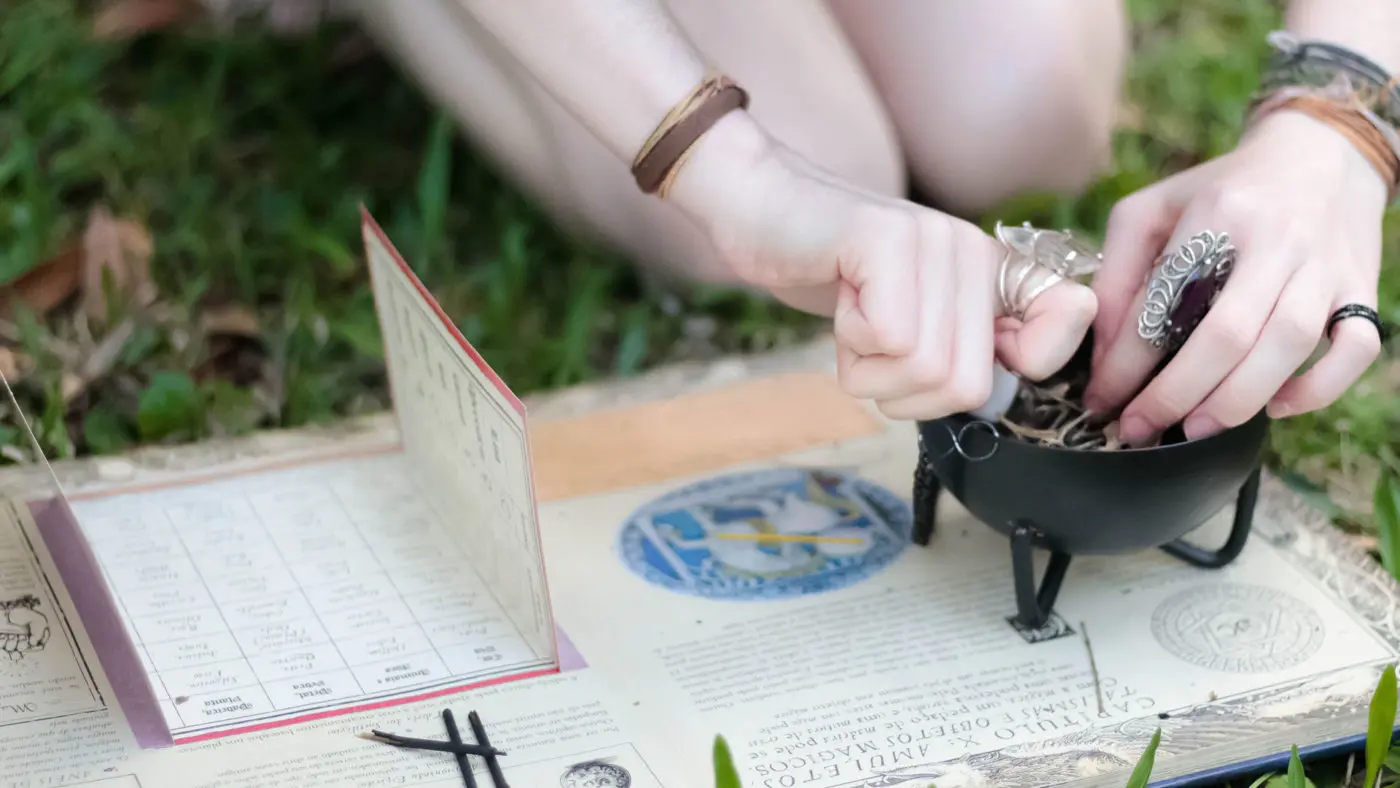 Where to shop for the Pagan Holidays 1 A person practicing a witchy divination or spellcasting ritual with various mystical accessories including a cauldron, tarot cards, and symbolic jewelry, set upon an outdoor earthy backdrop.