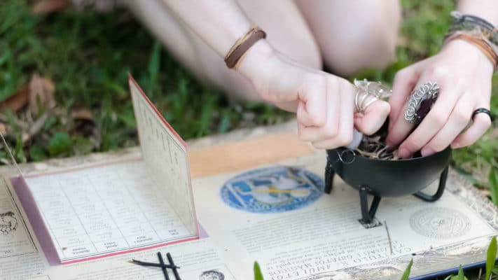 Where to shop for the Pagan Holidays 10 A person practicing a witchy divination or spellcasting ritual with various mystical accessories including a cauldron, tarot cards, and symbolic jewelry, set upon an outdoor earthy backdrop.