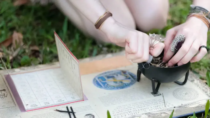 A person practicing a witchy divination or spellcasting ritual with various mystical accessories including a cauldron, tarot cards, and symbolic jewelry, set upon an outdoor earthy backdrop.