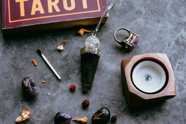 A mystical and witchy flat lay featuring tarot cards, an obsidian pendant, a candle holder, stones, and dried flowers on a textured dark surface, evoking a sense of divination and