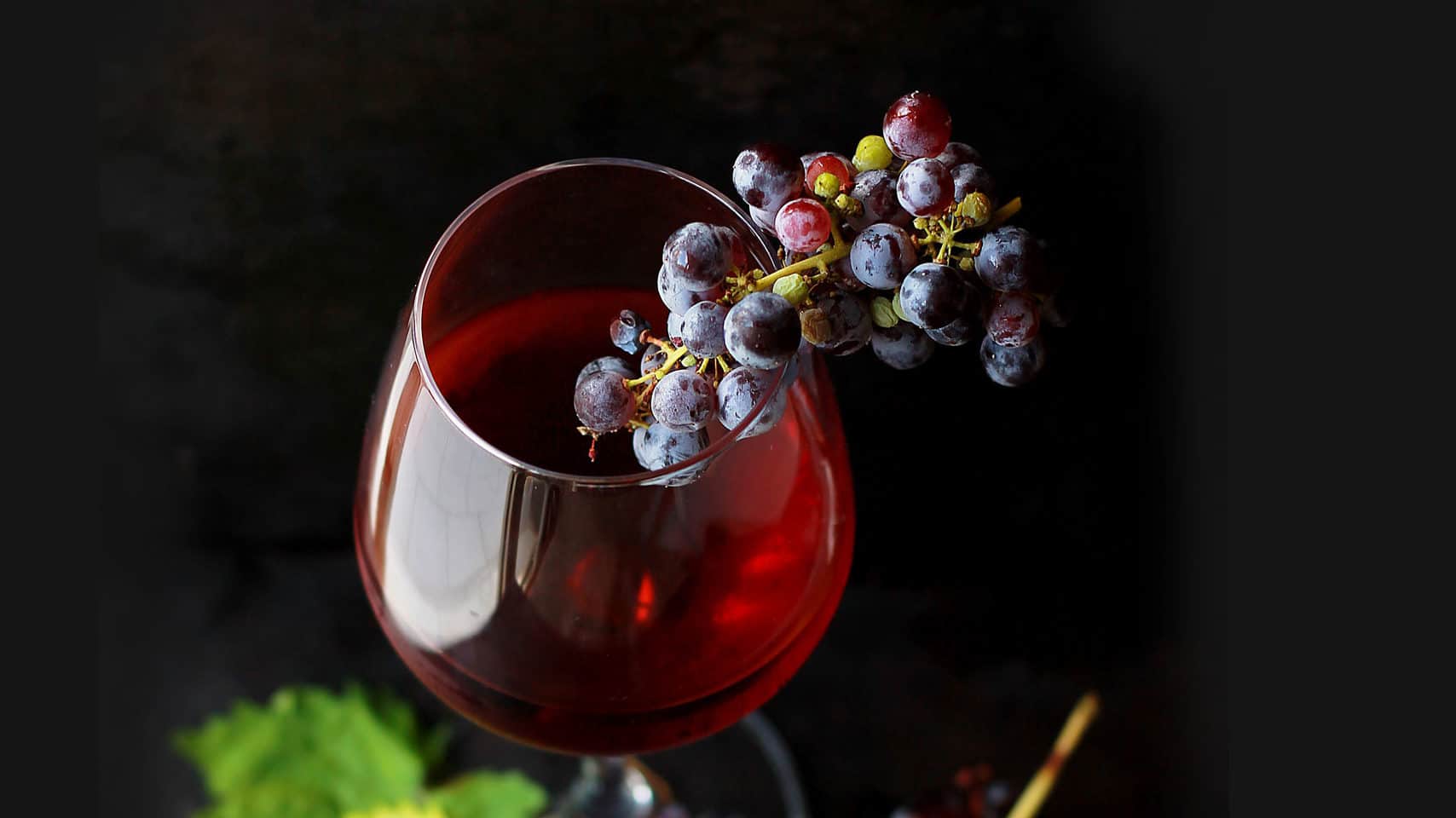 A glass of red wine elegantly accompanied by a cluster of grapes against a dark, witchy background.