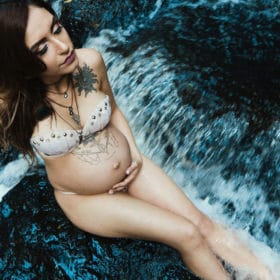 A pregnant individual poses elegantly on dark wet rocks with rushing water surrounding them, capturing a moment of calmness and connection with nature, embodying the witchy spirit of motherhood.