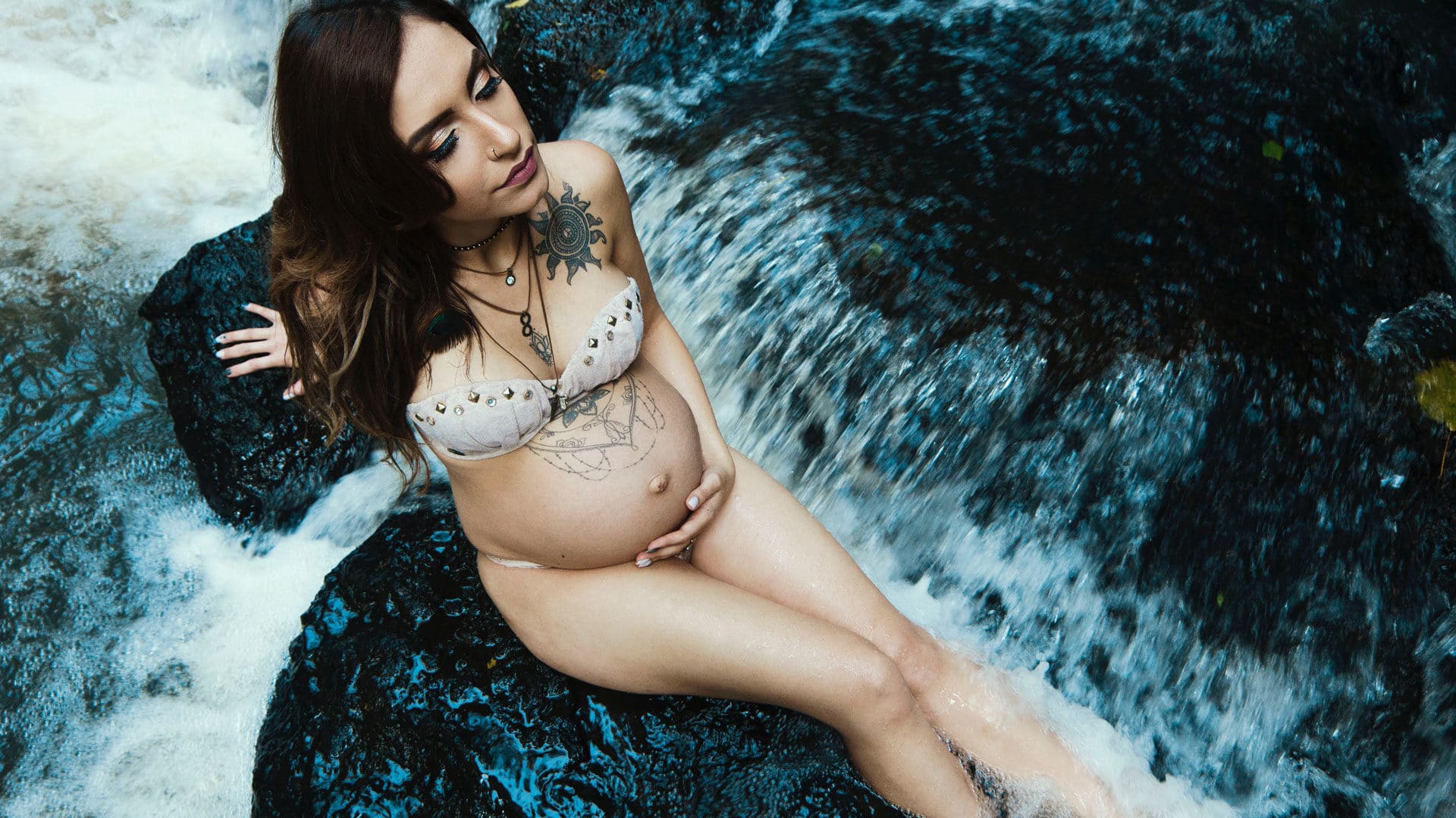 A pregnant individual poses elegantly on dark wet rocks with rushing water surrounding them, capturing a moment of calmness and connection with nature, embodying the witchy spirit of motherhood.