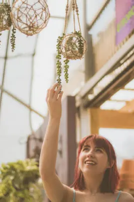 A young woman with red hair smiling as she gently touches a hanging decorative plant orb in a sunlit atrium, her presence exuding a witchy charm.
