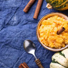 A bowl of warm, mashed pumpkin topped with cinnamon sticks on a vibrant blue cloth beside decorative gourds and witchy artifacts, evoking the cozy essence of autumn.