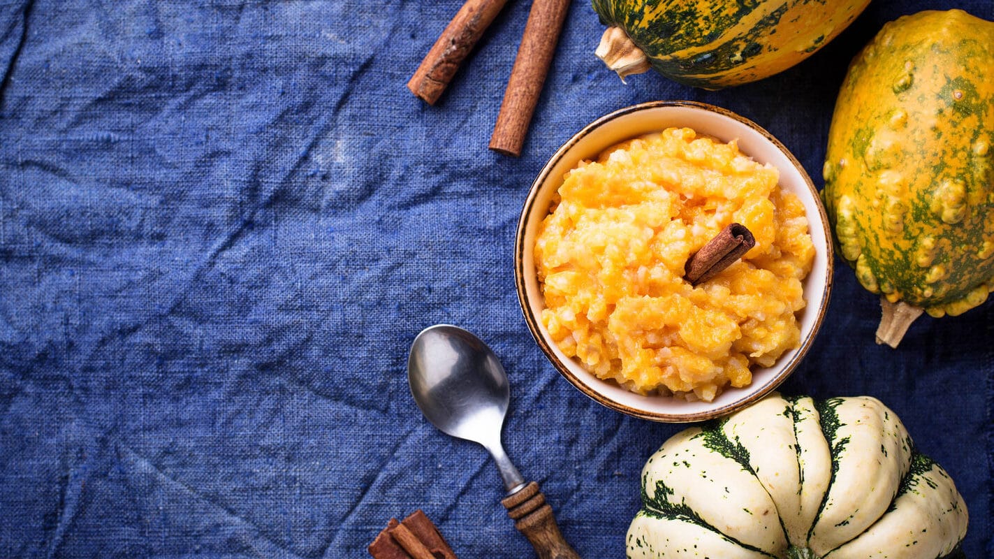 A bowl of warm, mashed pumpkin topped with cinnamon sticks on a vibrant blue cloth beside decorative gourds and witchy artifacts, evoking the cozy essence of autumn.