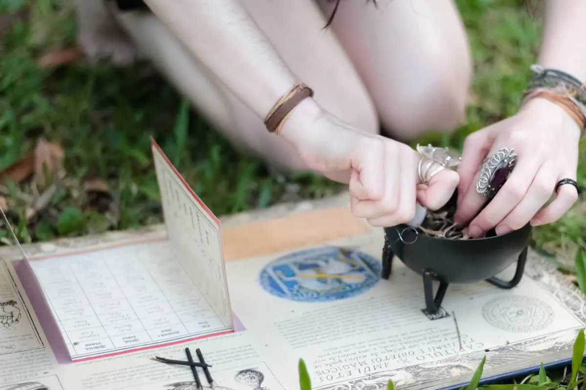A person practicing a ritual or spell with a cauldron and various tools on a grassy surface, possibly related to paganism or wicca, surrounded by metaphysical items.