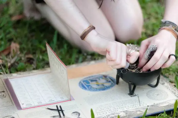 A person practicing a ritual or spell with a cauldron and various tools on a grassy surface, possibly related to paganism or wicca, surrounded by metaphysical items.