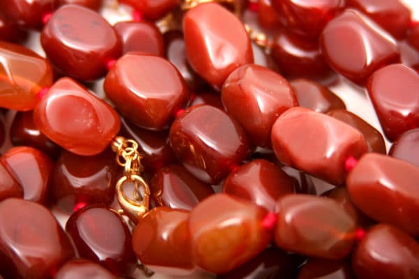 Magical Properties of Carnelian | Carnelian Materia Magicka 3 A close-up of polished red carnelian gemstones and beads with a gold clasp, creating a luxurious and vibrant background for a witchy or occult-themed metaphysical shop.