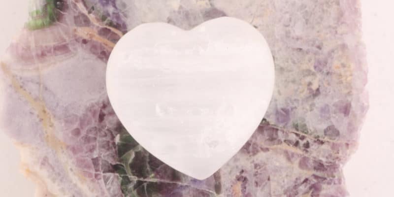 5 Ways to Use Selenite 2 A translucent heart-shaped stone rests upon a mottled purple and green mineral background, exuding a sense of serene beauty and spiritual harmony.