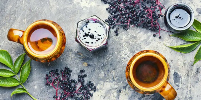 A top-down view of a rustic tea setting at a metaphysical shop featuring two mugs of tea alongside fresh and preserved elderberries, with green leaves adding a touch of nature.