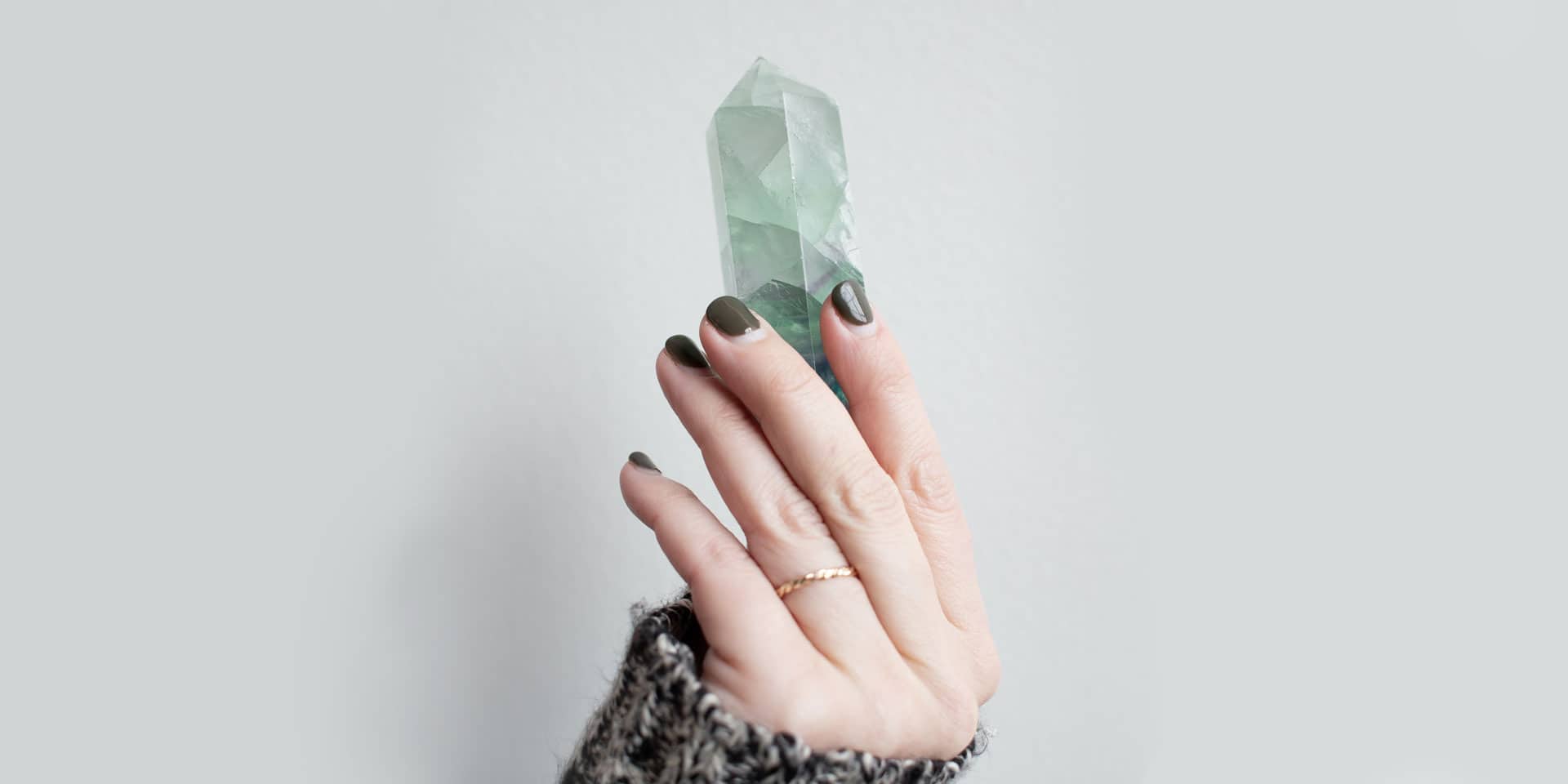 A person's hand with dark, witchy nail polish holding a large, translucent green crystal against a plain, light background.
