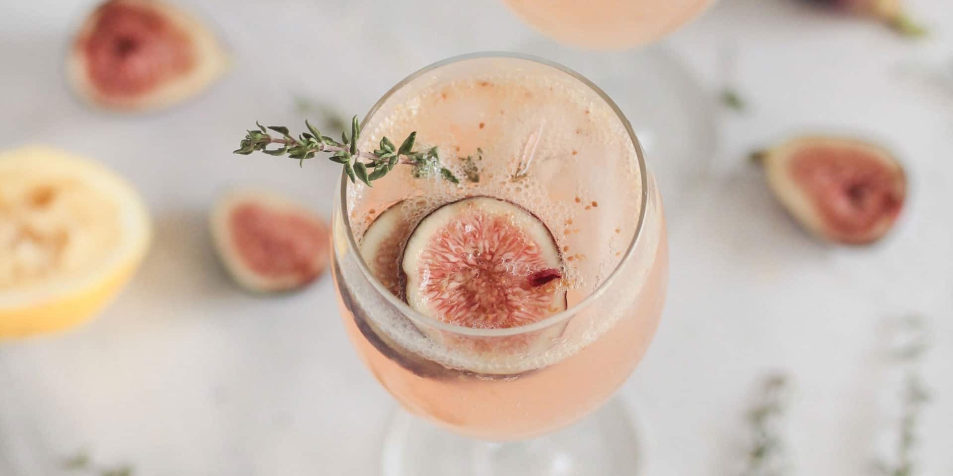 A refreshing fig cocktail garnished with a sprig of thyme and a slice of fig, with a subtle backdrop of fig halves and metaphysical herbs.
