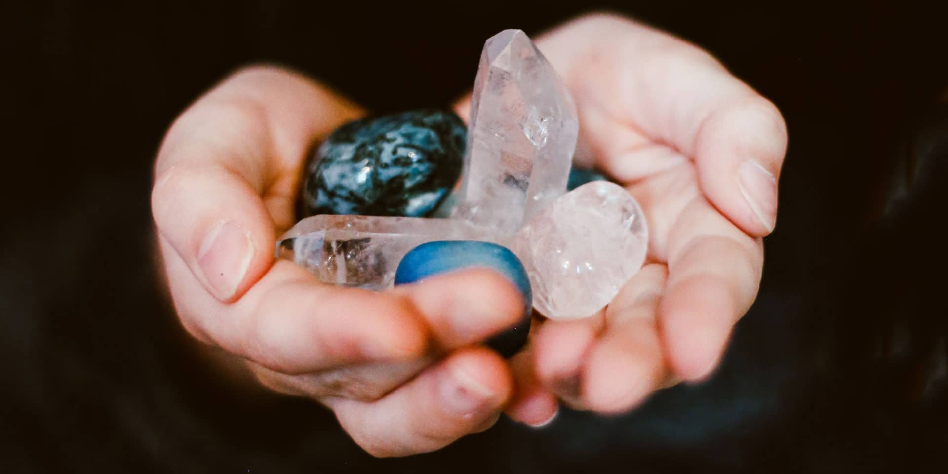 A close-up of a person's cupped hands gently holding a collection of various translucent and colored gemstones, radiating a witchy, metaphysical aura.
