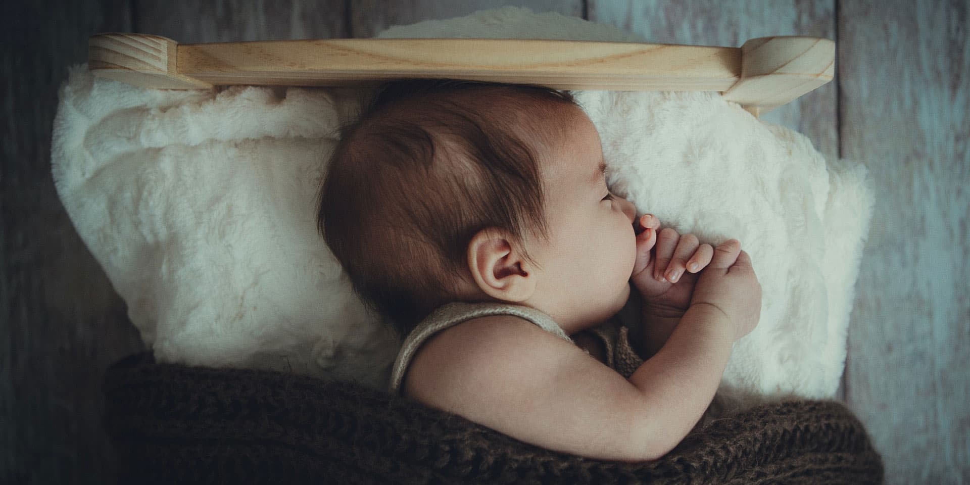 A peaceful infant asleep on a soft white blanket, cradled in a rustic wooden crib, with tiny hands gently resting by the face, conjuring a metaphysical serenity in the room.
