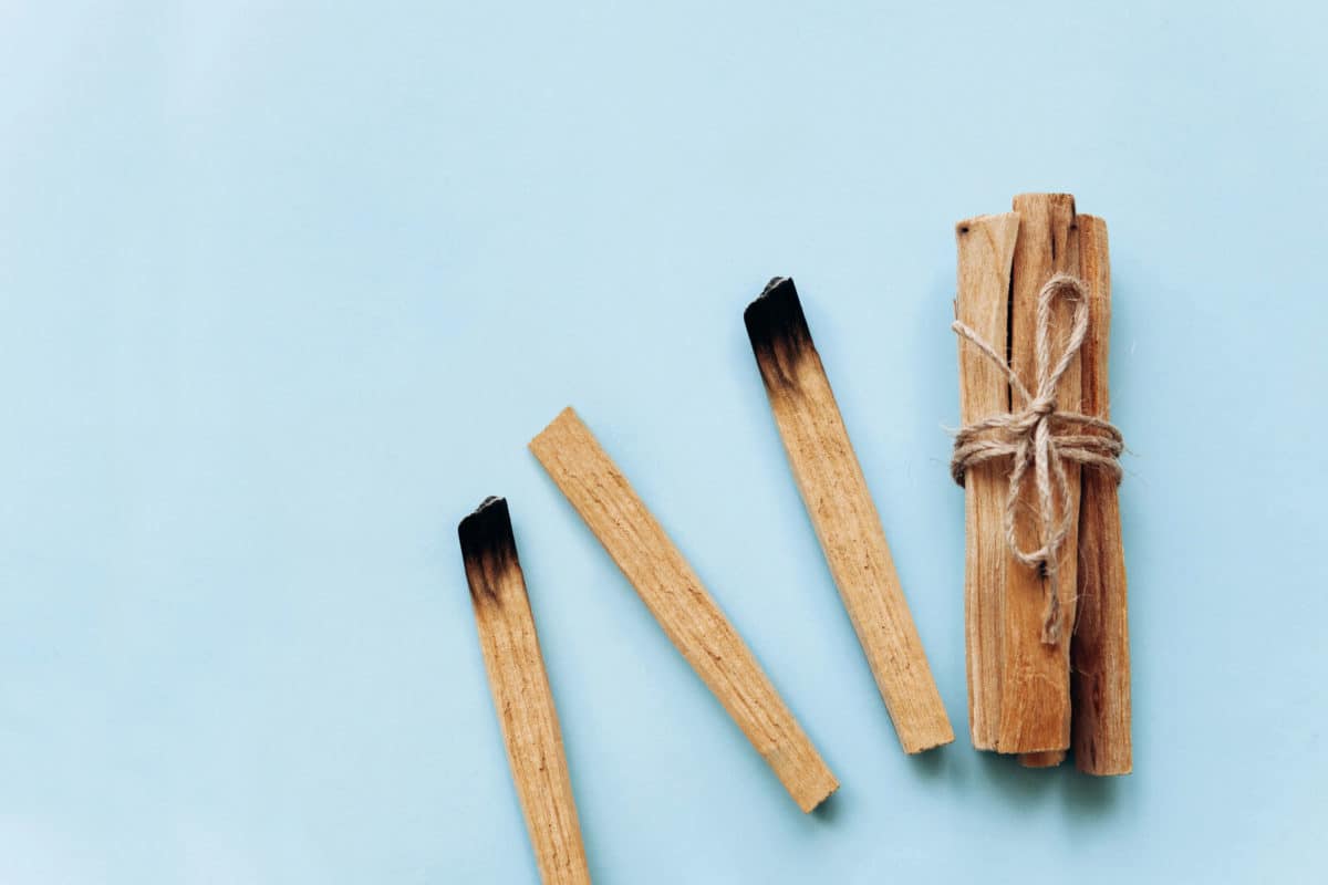 Three palo santo sticks, one bundle tied with twine, on a blue background, emanate a spiritual essence, with two of the sticks showing signs of being burnt on one end.