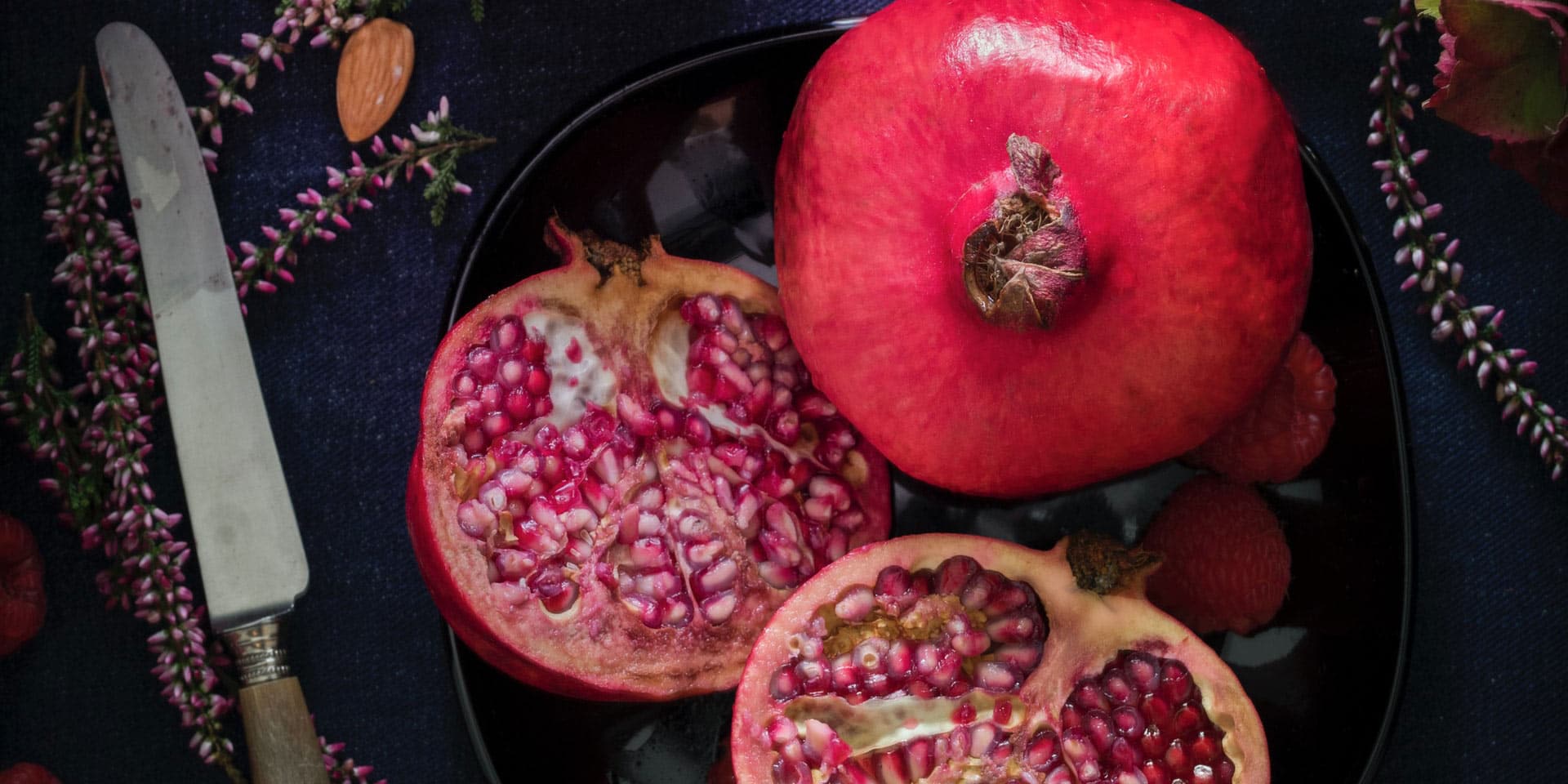 A whole and partially sliced pomegranate on a dark plate with a rustic knife to the side, surrounded by a sprinkling of almonds and hints of purple flowers, all set against a dark,