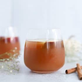 A refreshing glass of iced tea garnished with a cinnamon stick, evoking a witchy aura, with a soft-focus background featuring a second glass and delicate white flowers.