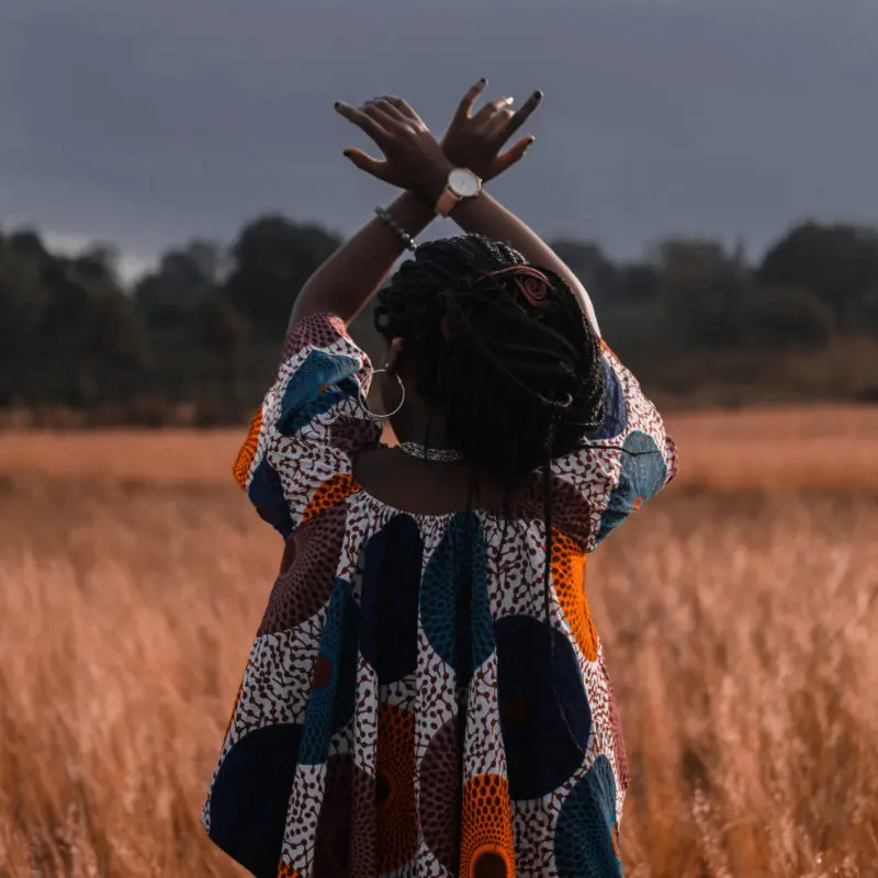 A person stands amidst tall grass, their arms raised towards the sky in a gesture of spiritual freedom or celebration, wearing a vibrantly patterned outfit against the backdrop of a subdued, dusky landscape.