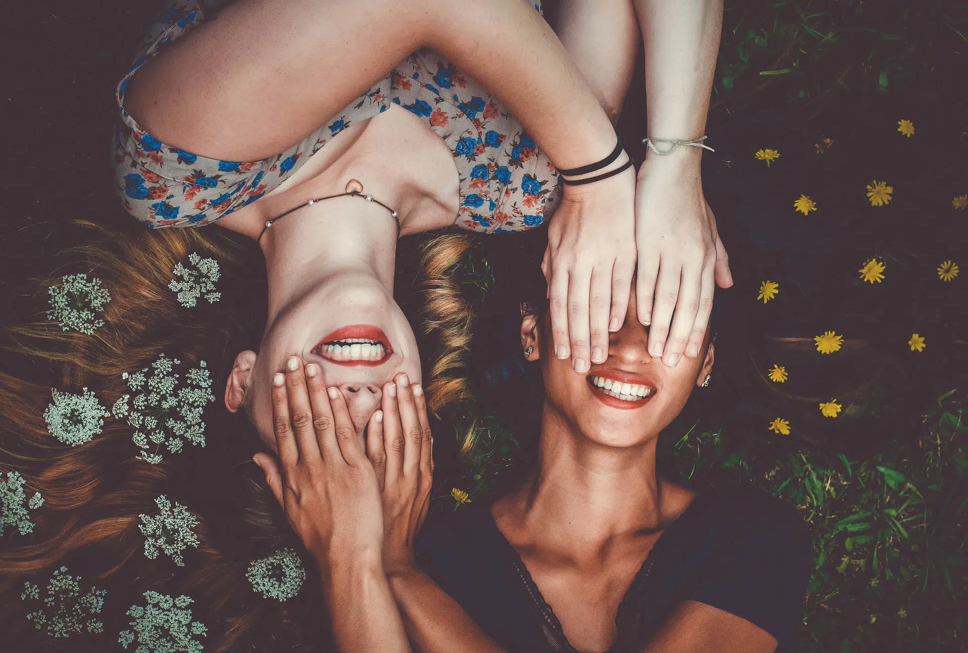Two friends lying on the grass, covering their eyes with their hands, smiling in a carefree moment surrounded by nature, embracing a metaphysical connection.