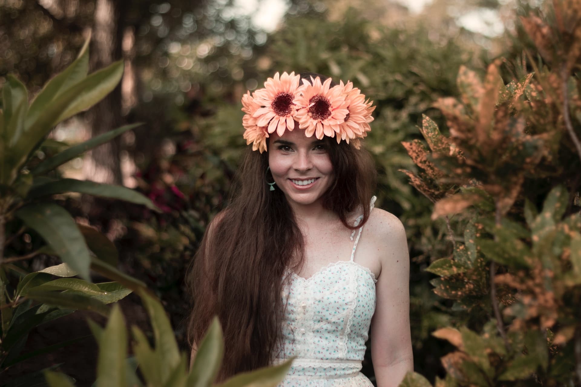 A smiling woman wearing a floral headpiece surrounded by lush greenery exudes a witchy aura.