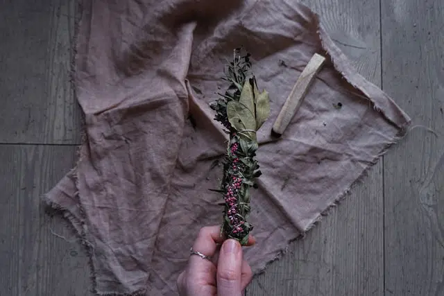A hand holding a bundle of dried herbs with a pink fabric and a piece of wood on a wooden surface for spiritual rituals.